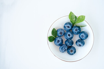 Fresh blueberries in a small white plate on a white background. Wild berry with green leaves, top view and copy cpace