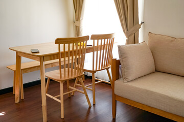 A clean, modern interior featuring a light wooden dining table with matching chairs, a beige upholstered sofa, and cream curtains allowing sunlight into the room.