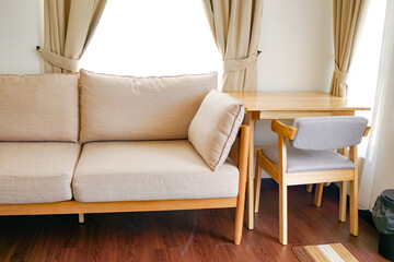 A cozy, well-organized living area featuring a beige fabric sofa, light wood dining table, cushioned chair, and beige curtains framing a sunlit window.