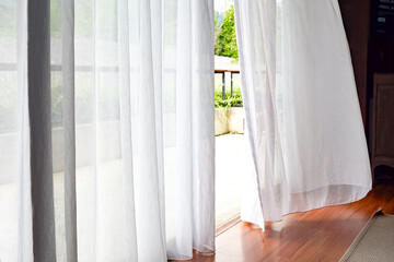 Bright indoor space with sheer white curtains billowing gently, revealing a wooden deck and lush greenery outside. Natural light floods the polished wood floor.