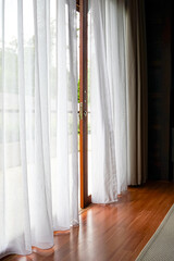 Bright indoor space with sheer white curtains billowing gently, revealing a wooden deck and lush greenery outside. Natural light floods the polished wood floor.