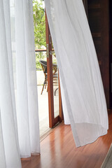 Bright indoor space with sheer white curtains billowing gently, revealing a wooden deck and lush greenery outside. Natural light floods the polished wood floor.