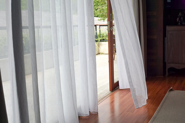 Bright indoor space with sheer white curtains billowing gently, revealing a wooden deck and lush greenery outside. Natural light floods the polished wood floor. © JedRT
