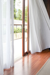 Bright indoor space with sheer white curtains billowing gently, revealing a wooden deck and lush greenery outside. Natural light floods the polished wood floor.