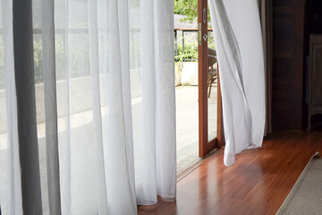 Bright indoor space with sheer white curtains billowing gently, revealing a wooden deck and lush greenery outside. Natural light floods the polished wood floor.