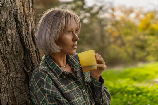 Woman nature coffee enjoying morning hot beverage leaning on a rustic tree trunk outdoors