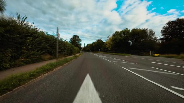 Car heading along quiet suburban road under cloudy blue sky surrounded by trees POV. Automobile proceeding through open countryside intersection bordered by greenery first person view. Vehicle