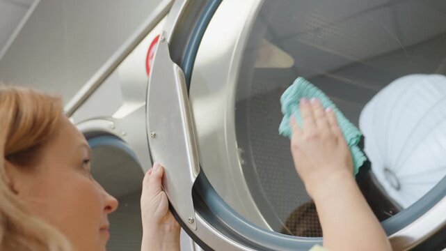 Upside down view of worker hands wiping surface of industrial washing machine with wet towel, focus on cleaning motion showing reflection on glass door and details of metallic laundry equipment