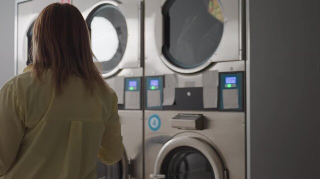 Back view cleaner woman carrying laundry basket through dim laundromat, industrial washers running with fabrics visible behind glass, stainless panels, routine operation, indoor workplace