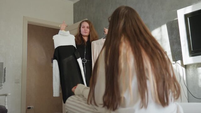 Lower view of roommates interacting indoors as one seated on bed points towards chosen outfit while other stands holding two dresses on hangers, showing casual fashion decision