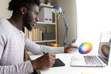 Black male graphic designer working at home using laptop and graphic tablet.