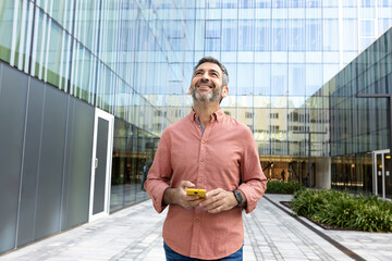 Happy mature man walking in the city holding mobile phone looking away. Copy space.