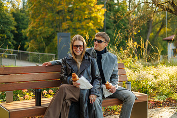 Fototapeta premium Joyful man and woman sit close together on a park bench, holding croissants and coffee cups, dressed stylishly for fall. Surrounded by golden foliage. No visible logos