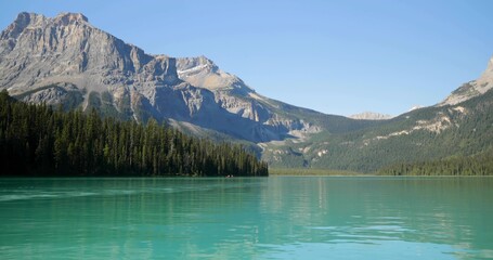 Showing rugged mountain peak rising above evergreen tree line at turquoise lake with snow patches