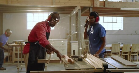 Two carpenters with safety goggles and ear protection cutting wooden board on table saw in workshop