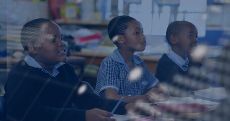 Writing students holding pencils at desks in classroom, with textbooks notebooks and bookshelves