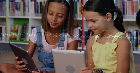 Reading two schoolgirls crosslegged using tablets on library carpet, with bookshelves backdrop