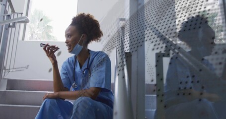 Sitting nurse in scrubs speaking into smartphone in stairwell, with surgical mask and stethoscope