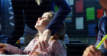 Pointing teacher leaning over girl student desk in classroom, with worksheets and pencils