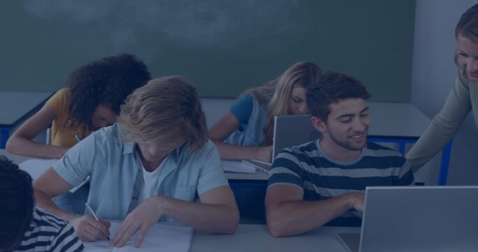 Instructor helping students at desks in classroom with laptops, notebooks and chalkboard background
