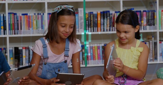 Sitting two girls in denim overalls, yellow bow top reading tablets in library, with bookshelves
