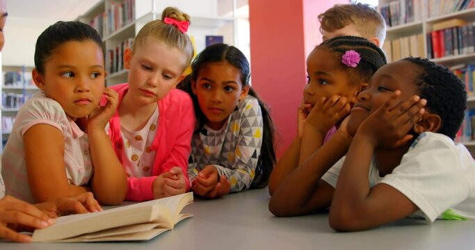 Leaning classmates reading open book at gray table in library, with bookshelves and red column