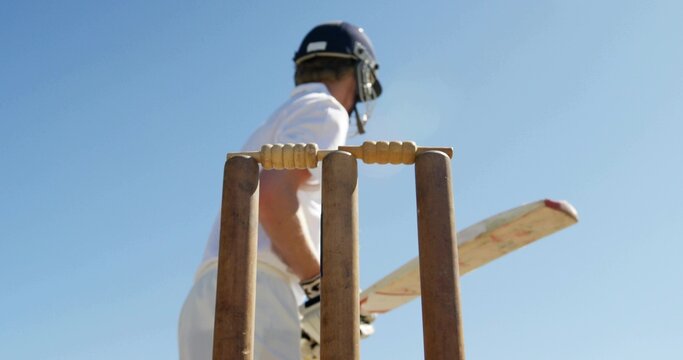 Framing cricket wicket with stumps on cricket pitch, batsman wearing white kit, helmet holding bat