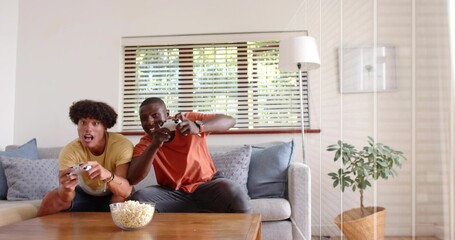 Playing two friends sitting on light-gray sofa in living room, holding controllers and popcorn bowl