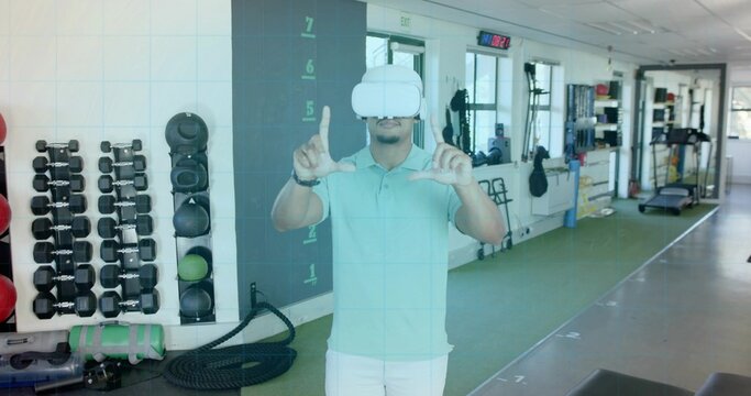 Framing man wearing white VR headset and teal polo forming rectangle gesture in gym with dumbbells - Powered by Adobe
