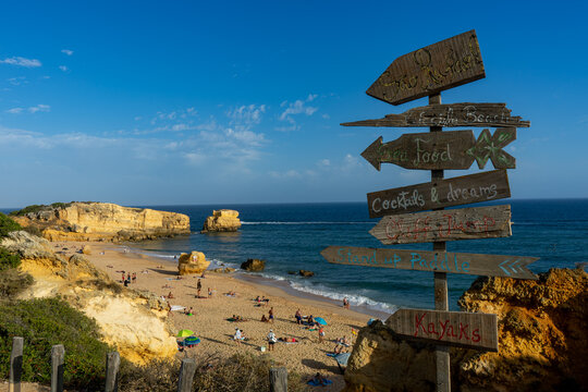 A rustic wooden signpost at S&atilde;o Rafael Beach, Portugal, points towards the sea and late-afternoon sun with playful messages for seafood, cocktails and seaside adventures