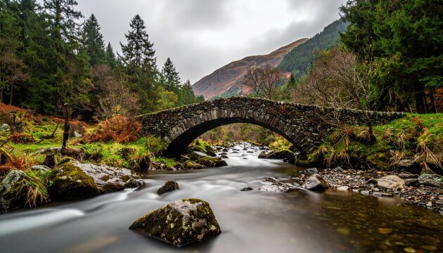 Stone Arch Bridge Over A Rushing River Amidst Autumn Forest Trees And Mountains With A Cloudy Sky Overhead - Powered by Adobe
