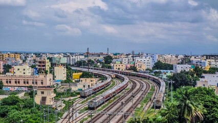 Curved railway tracks lead into the heart of Tirupati city, surrounded by colorful residential blocks and lush greenery. The cloudy sky adds a calm monsoon mood to this vibrant and fast-growing city