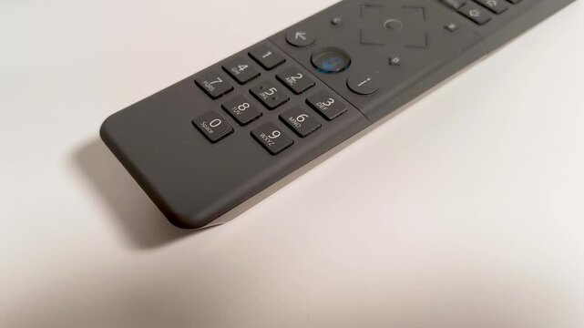 Modern dark gray remote control with illuminated microphone button and numeric keypad resting on a clean white surface, ready for entertainment or smart home interaction