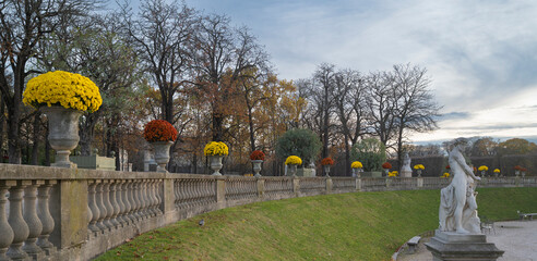 Visitors stroll through the Luxembourg Gardens in Paris, admiring autumn flowers in planters. The scene features stone statues. High quality photo