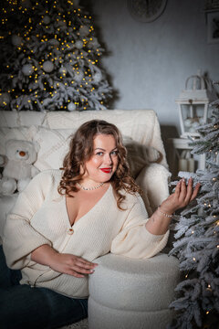 A portrait of a beautiful,chubby  woman with a sweet smile against a New Year's backdrop. A buxom woman in a white knitted sweater with an open neckline poses against a backdrop of Christmas lights.