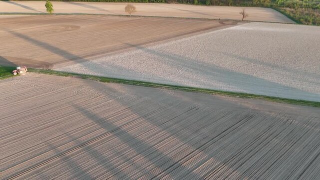 Drone-captured agricultural landscape reveals straight furrows and sharp parcel boundaries, illuminated by long evening shadows fromt rees and a lone red tractor stopped by.