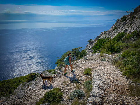 Sporty young woman walks uphill along a rocky coastal trail with her dog under bright summer sun. Outdoor exploration, companionship, and beauty of Mediterranean wilderness high above shimmering sea.