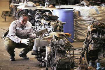 Black female technician check used car damaged engine at scrapyard warehouse recycle area part. African American engineer inspecting rusty oily auto motor old spare part in junkyard for reuse service
