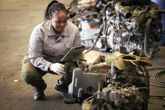 Black female technician check used car damaged engine at scrapyard warehouse recycle area part. African American engineer inspecting rusty oily auto motor old spare part in junkyard for reuse service
