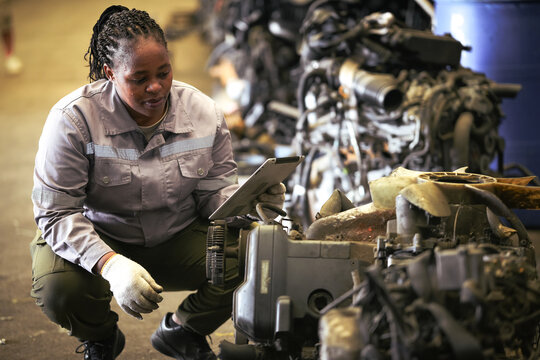 Black female technician check used car damaged engine at scrapyard warehouse recycle area part. African American engineer inspecting rusty oily auto motor old spare part in junkyard for reuse service