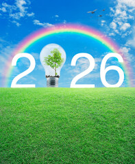 Naklejka premium 2026 white text and light bulb with tree inside on green grass field over blue sky, white clouds and rainbow, Happy new year 2026 ecological cover concept
