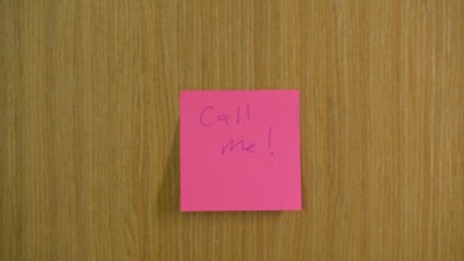 Vibrant pink sticky note attached to wooden door with handwritten 'Call me!'. Focused close-up shot conveying urgent communication