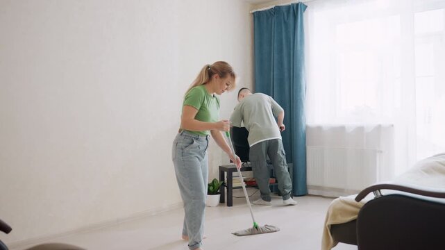 Family cleans living room as woman wipes floor with mop while son dusts television, sunlight shines through bright window, teamwork and cooperation transform household chores