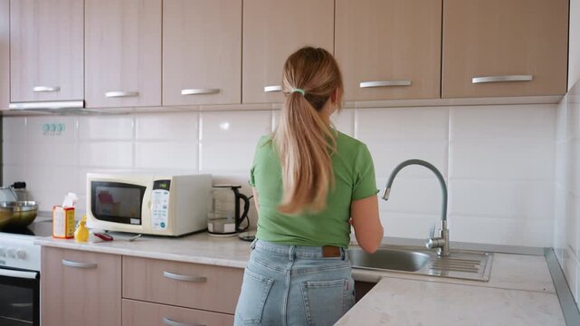 Back view of lady tidying modern kitchen as she puts black cup into upper cabinet, maintaining order while surrounded by microwave, sink, stove, and utensils in daily household routine