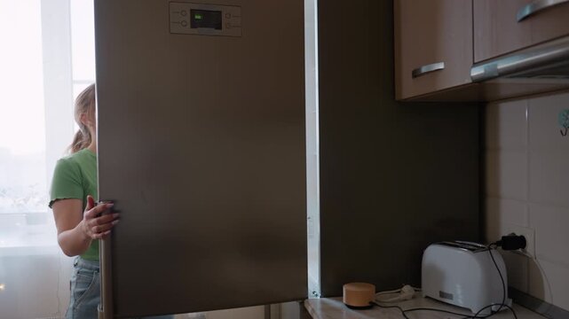 Side view of woman in green shirt retrieving eggs from refrigerator in kitchen, preparing ingredients for cooking while standing near counter and cabinets in bright domestic interior