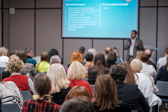 Audience professionals attend live presentation in bright conference room. Focused attendees watch speaker share insights while slides illuminate background