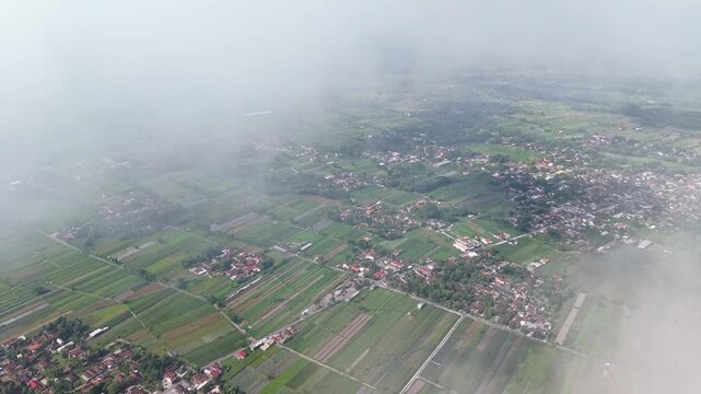 Aerial view of agricultural land with geometric patterns of fields and scattered houses under a blanket of mist, Klaten Regency, Central Java, Indonesia.