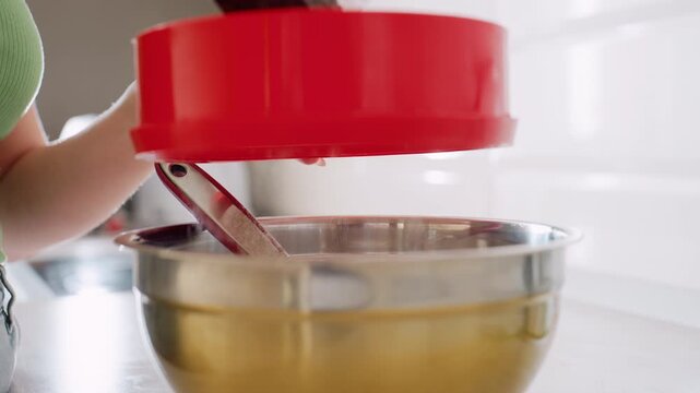 Close up of chef in green top using red sifter to filter flour into stainless bowl during baking preparation, flour dust falling smoothly while mixing utensil rests inside bowl on kitchen counter