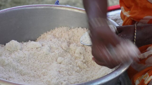 Elderly woman mixing coarse idli batter in large metal basin at outdoor street food stall