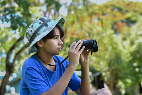 A young boy wearing a blue t-shirt and a camouflage cap uses a pair of binoculars to look off-camera, standing amidst dense greenery in a forest or park setting.
